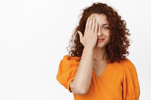 Schönheit und Wohlbefinden der Frauen. Junge Frau mit rotem, lockigem Haar, Gesichtshälfte, mit einer Augenseite schauend und selbstbewusst lächelnd, auf Weiß stehend.