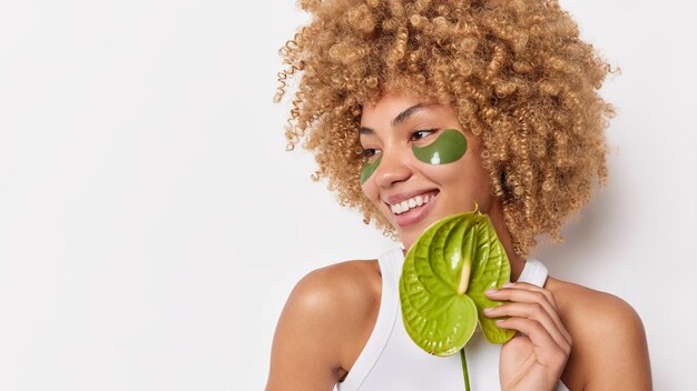 Schönheit wirksame Behandlung. Positive Frau mit blonden lockigen Haaren hält Calla-Blume sieht glücklich weg, trägt grüne Hydrogel-Patches auf, die in T-Shirt gekleidet sind, isoliert auf weißem Hintergrund, leerer Kopienraum