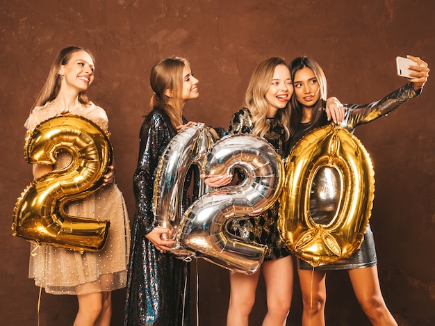 Schönheiten, die neues Jahr feiern Glückliche herrliche Mädchen in den stilvollen sexy Parteikleidern, die Gold- und Silber-2020-Ballone, Spaß an Silvesterabend-Party habend halten Selfie oder Video für instagram machen