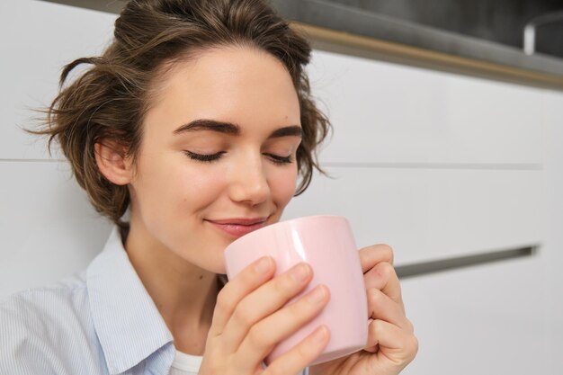 Schönheits- und Frauenkonzept. Entspannte junge Frau genießt ihre Tasse Tee. Mädchen sitzt witzig auf dem Boden in der Küche