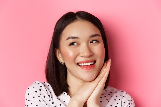 Schönheits- und Hautpflegekonzept. Kopfschuss einer entzückenden und verträumten asiatischen Frau, die nach links schaut, lächelt und sich vorstellt, vor rosa Hintergrund steht