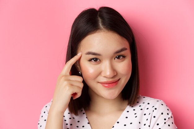 Schönheits- und Hautpflegekonzept. Kopfschuss einer intelligenten asiatischen Frau, die auf den Kopf zeigt und schlau lächelt, zum Nachdenken auffordert und über rosafarbenem Hintergrund steht