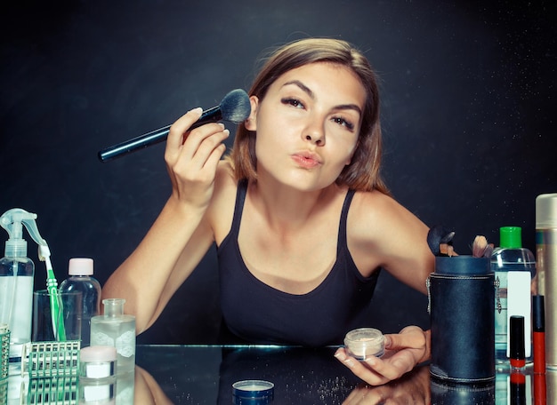 Kostenloses Foto schönheitsfrau, die make-up anwendet. schönes mädchen, das in den spiegel schaut und kosmetik mit einem großen pinsel anwendet. kaukasisches modell im studio