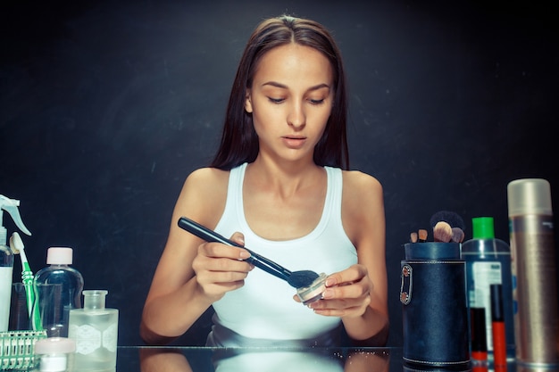 Schönheitsfrau, die make-up anwendet. schönes mädchen, das in den spiegel schaut und kosmetik mit einem großen pinsel anwendet. morgen, make-up und menschliches gefühlskonzept