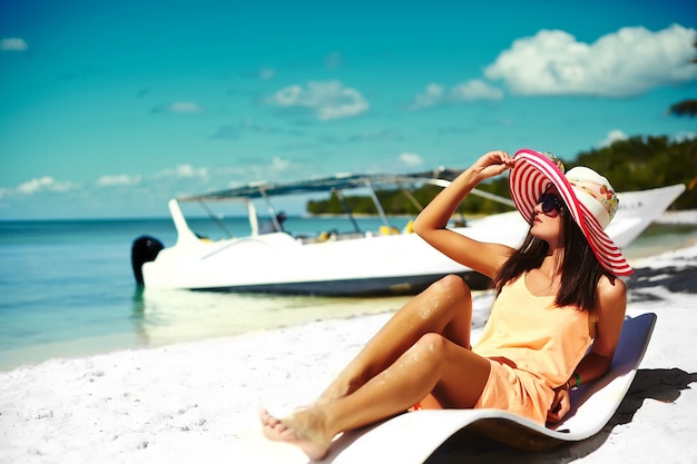 Schönheitsmodell, das auf dem Strandstuhl im weißen Bikini im bunten sunhat hinter blauem Sommerwasserozean ein Sonnenbad nimmt