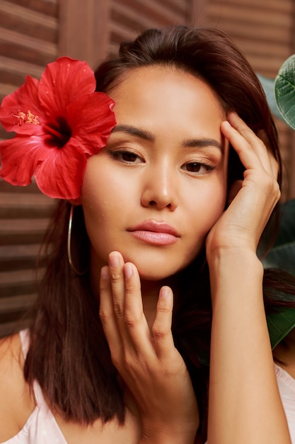 Schönheitsporträt der asiatischen Frau mit perfekter Haut und Hibiskusblume in den Haaren, die über Holzwand aufwerfen