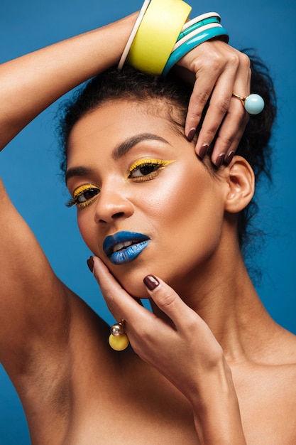 Schönheitsporträt der jungen Frau des attraktiven Afroamerikaners mit Modemake-up und -Zubehör auf der Handaufstellung lokalisiert, über blauer Wand