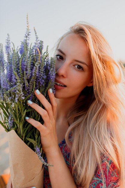 Schönheitsporträt der reizenden blonden Frau mit Blumenstrauß des Lavendels, der Kamera betrachtet. Perfekte Haut. Natürliches Make-up.