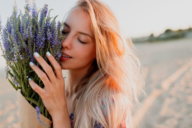 Schönheitsporträt der romantischen blonden Frau mit dem Blumenstrauß des Lavendels, der Kamera betrachtet. Perfekte Haut. Natürliches Make-up.