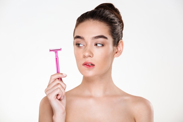 Schönheitsporträt der weiblichen Frau mit der weichen sauberen Haut, die rosa Rasiermesser in ihrer Hand sich vorbereitet für Körperbehandlung betrachtet