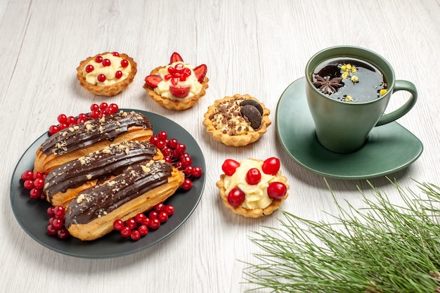 Schokoladen-Eclairs und Johannisbeeren von unten auf dem grauen Teller, umgeben von Torten, einer Tasse Tee und Kiefernblättern auf dem weißen Holztisch