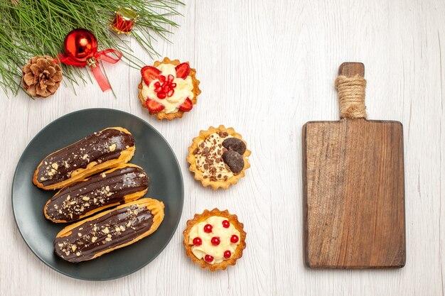 Schokoladen-Eclairs von oben auf dem grauen Teller tört die Kiefernblätter mit Weihnachtsspielzeug links und einem Schneidebrett rechts vom weißen Holzboden