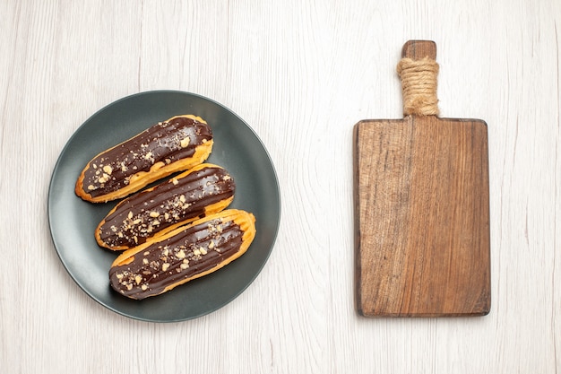 Schokoladen-Eclairs von oben auf dem grauen Teller und ein Schneidebrett auf dem weißen Holzgrund
