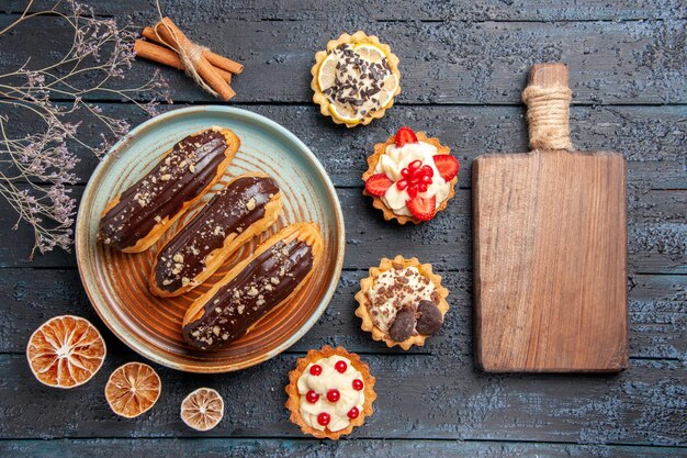 Schokoladen-Eclairs von oben auf einem ovalen Teller, umgeben von getrockneten Zitronentörtchen und Zimt und einem Schneidebrett auf dem dunklen Holztisch