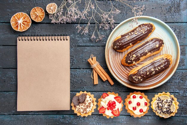 Schokoladen-Eclairs von oben auf ovalen Tellertörtchen mit zimtgetrockneten Orangen und einem Notizbuch auf dem dunklen Holztisch