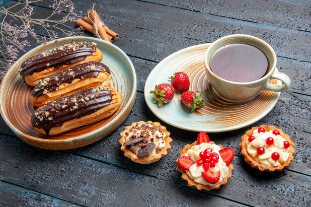 Schokoladen-Eclairs von unten auf ovalem Teller eine Tasse Tee und Erdbeeren auf Untertassen-Törtchen und Zimt auf dem dunklen Holztisch