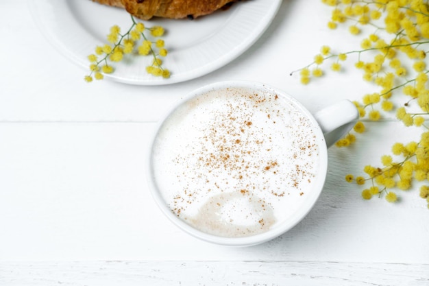 Schokoladencroissant und Cappuccino mit Mimosenblumen auf einem weißen Tisch Frühlingskonzept zum Muttertag