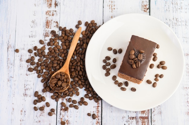 Schokoladenkuchen auf einem weißen Teller und Kaffeebohnen auf einem Holzlöffel auf einem Holztisch.