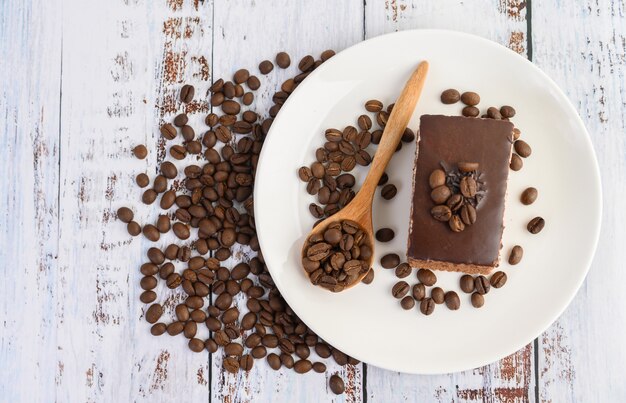 Schokoladenkuchen auf einem weißen Teller und Kaffeebohnen auf einem Holzlöffel auf einem Holztisch.