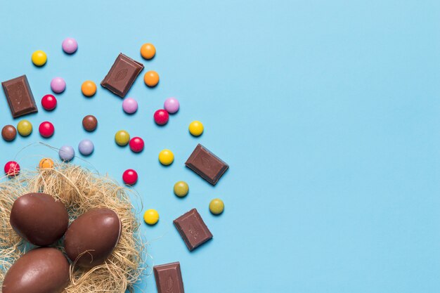 Schokoladenostereier im Nest verziert mit Edelsteinsüßigkeiten und Schokoladenstücken auf blauem Hintergrund