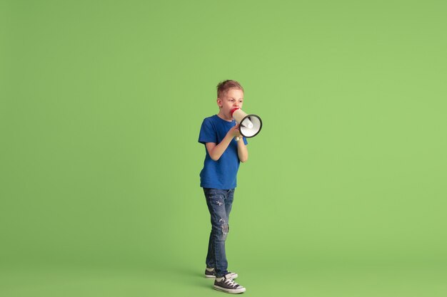 Schreien, rufen. Fröhlicher Junge, der auf grüner Wand spielt und Spaß hat. Kaukasisches Kind in hellem Tuch sieht verspielt aus und lächelt. Konzept der Bildung, Kindheit, Emotionen, Gesichtsausdruck.