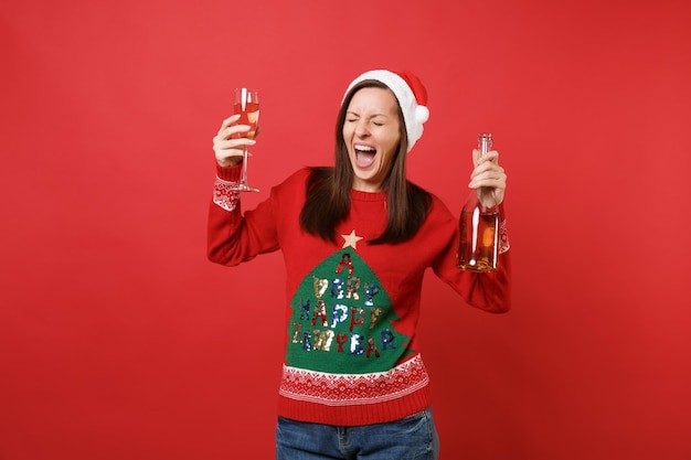 Schreiendes junges santa-mädchen in weihnachtsmütze, das die augen geschlossen hält und eine glasflasche champagner auf rotem hintergrund hält. frohes neues jahr 2019 feier urlaub party konzept. kopieren sie platz.