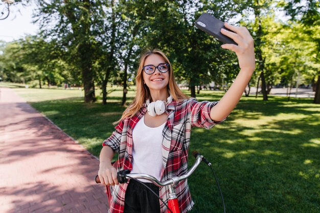 Schüchterne blonde Frau in der Brille, die Telefon für Selfie in guten Sommertag verwendet. Hübsches kaukasisches Mädchen, das mit rotem Fahrrad aufwirft.