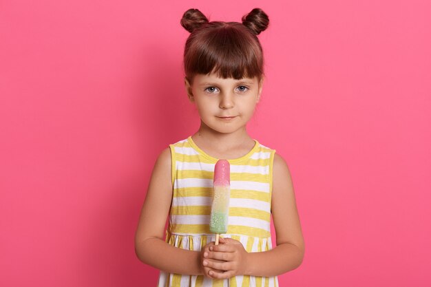 Schüchternes charmantes Mädchen mit Sorbet in den Händen, kleidet Sommeroutfit, hat zwei Knoten, die isoliert auf rosa stehen