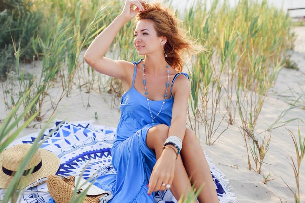 Schüchternes Mädchen mit perfekter Bräunungshaut, die auf sonnigem Strand im trendigen blauen Kleid auf Sand sitzend aufwirft. Windige Haare. Abendsonnenlicht.