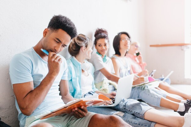 Schüler sitzen mit Laptops auf dem Boden und machen gemeinsam ihre Hausaufgaben. Schwarzer Junge mit der trendigen Frisur, die über schwierige Aufgabe nachdenkt und Stift in der Hand hält.