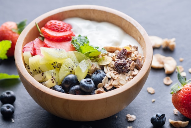 Schüssel Hafergranola mit Joghurt, frischen Blaubeeren, Erdbeeren, Kiwi-Minze und Nüssen für ein gesundes Frühstück, gesundes Frühstücksmenükonzept. Auf dem schwarzen Felsen