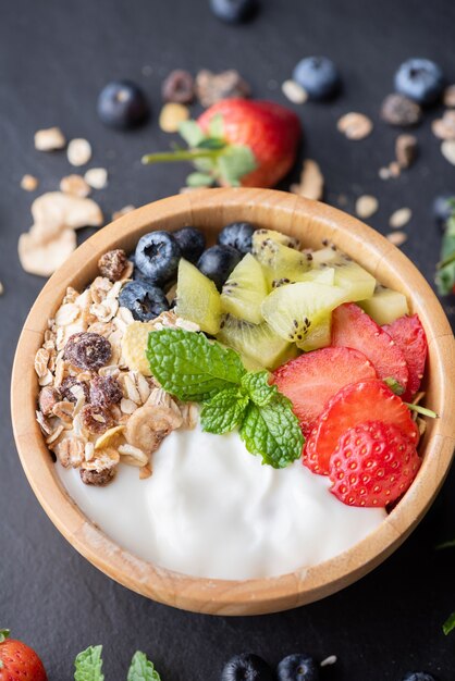 Schüssel Hafergranola mit Joghurt, frischen Blaubeeren, Erdbeeren, Kiwi-Minze und Nüssen für ein gesundes Frühstück, gesundes Frühstücksmenükonzept. Auf dem schwarzen Felsen