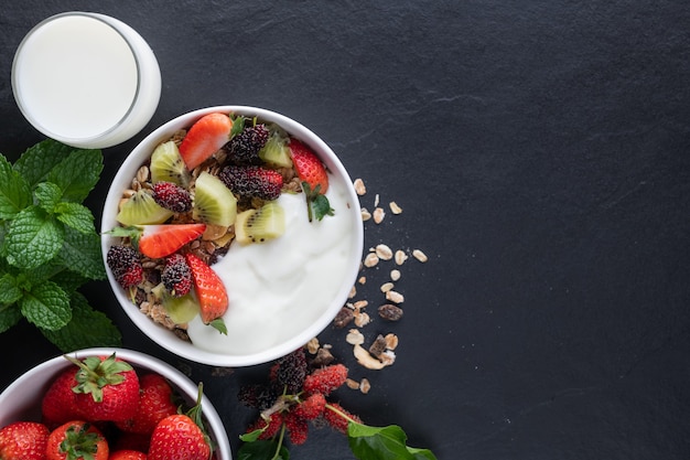 Schüssel Hafergranola mit Joghurt, frischer Maulbeere, Erdbeeren, Kiwi-Minze und Nüssen auf dem schwarzen Felsenbrett für gesundes Frühstück, Draufsicht, Kopienraum, flache Lage. Gesundes Frühstücksmenükonzept.