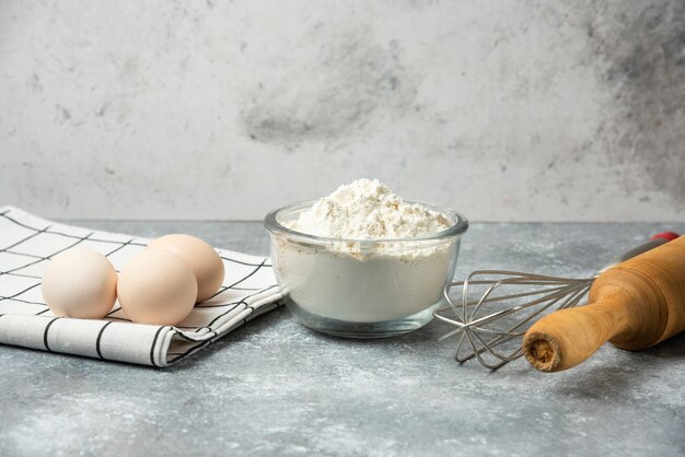 Schüssel Mehl, Eier und Küchenutensilien auf Marmortisch.