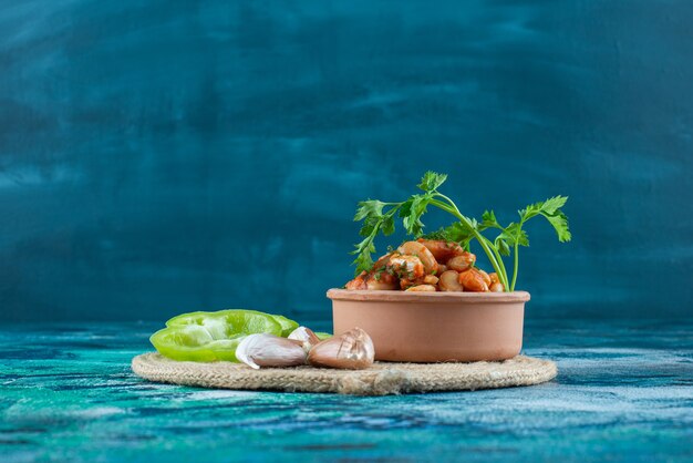 Schüssel mit gebackenen Bohnen mit Petersilie neben Knoblauch und Pfeffer auf einem Untersetzer, auf dem blauen Tisch.