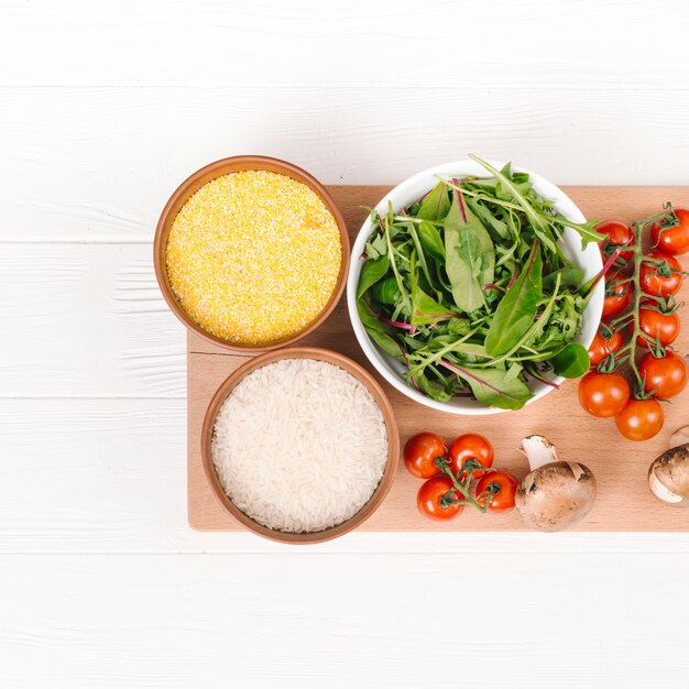 Kostenloses Foto schüssel mit polenta; reiskörner; mangold; pilz- und kirschtomaten auf schneidebrett über der weißen planke