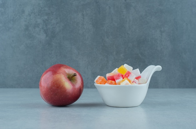 Schüssel Süßigkeiten und ein frischer Apfel auf Marmor.