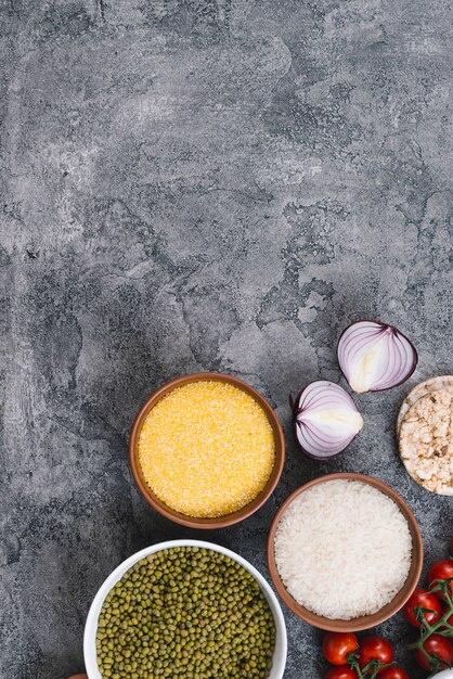 Schüsseln mit Reiskörnern; Mungobohnen; Polenta; Zwiebel; Kirschtomaten und Puffreiskuchen auf konkretem Hintergrund