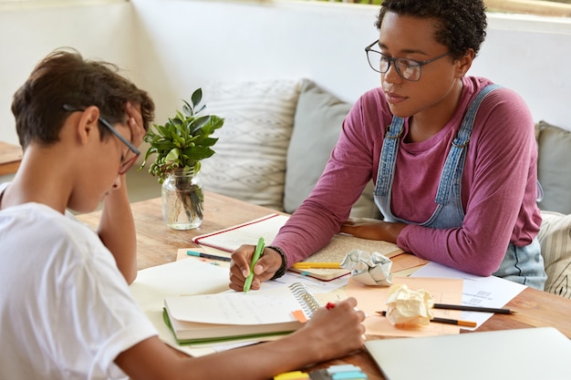 Schulbildung und Nachhilfekonzept. Horizontale Aufnahme der schwarzen klugen Afroamerikanerfrau antwortet auf eine Frage des Schülers, der Kopfschmerzen hat und Flipchart oder Diagramm nicht verstehen kann