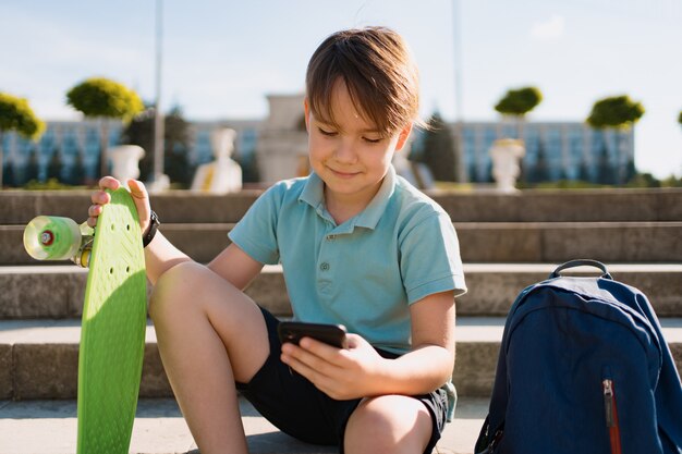 Schuljunge im blauen Poloshirt, das auf der Treppe mit einem blauen Rucksack und grünem Pennyboard unter Verwendung des Smartphones sitzt