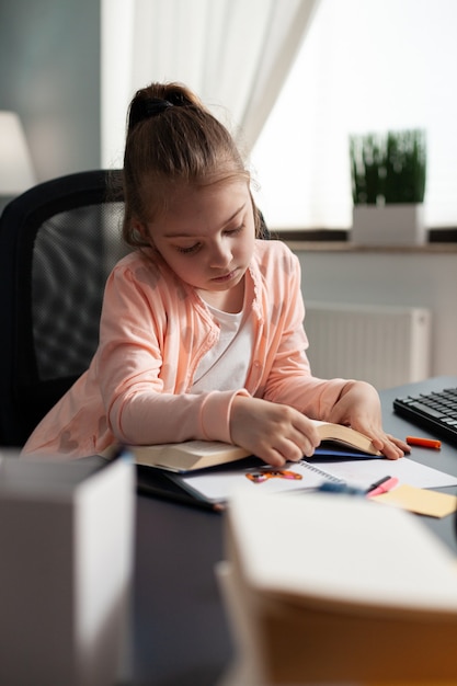 Schulkind sitzt am Schreibtisch im Wohnzimmer und hält Hausaufgabenheft