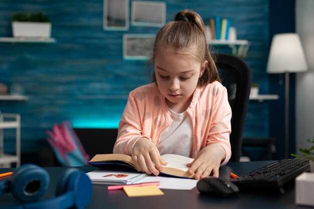 Schulkind sitzt am Schreibtisch im Wohnzimmer und hält Schulbuch