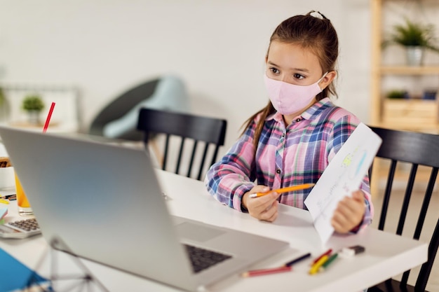 Schulmädchen mit Gesichtsmaske beim Online-Videounterricht zu Hause während der Sperrung des Coronavirus