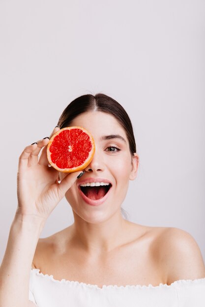 Schuss des Modells mit grauen Augen auf isolierter Wand. Positives Mädchen ohne Make-up bedeckt Auge mit halber Grapefruit.