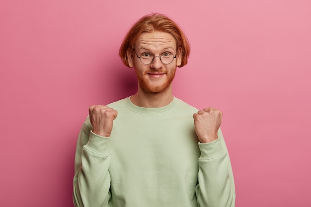 Kostenloses Foto schuss von erleichtertem ingwer der kaukasische mann ballt die fäuste, erwartet, dass etwas gutes passiert ist, trägt eine runde brille und einen pullover, isoliert über der rosa pastellwand. körpersprachenkonzept