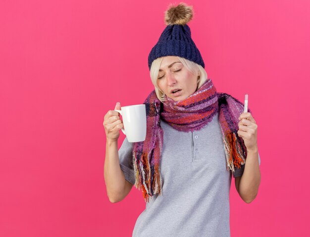 Schwache junge blonde kranke slawische Frau, die Wintermütze und Schal trägt, hält Tasse