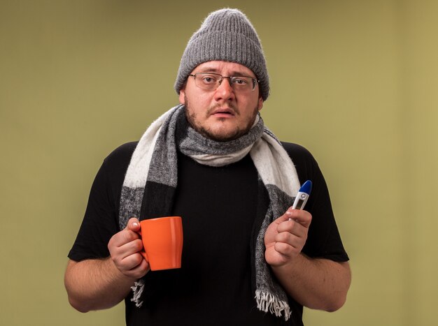 Schwacher Blick in die Kamera kranker Mann mittleren Alters mit Wintermütze und Schal, der eine Tasse Tee mit Thermometer hält