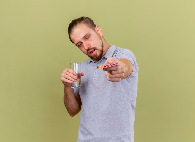 Schwacher junger hübscher slawischer kranker Mann, der Packung der Kapseln und Glas des Wassers hält, die Packung der Kapseln in Richtung Kamera mit geschlossenen Augen strecken, lokalisiert auf olivgrünem Hintergrund mit Kopienraum