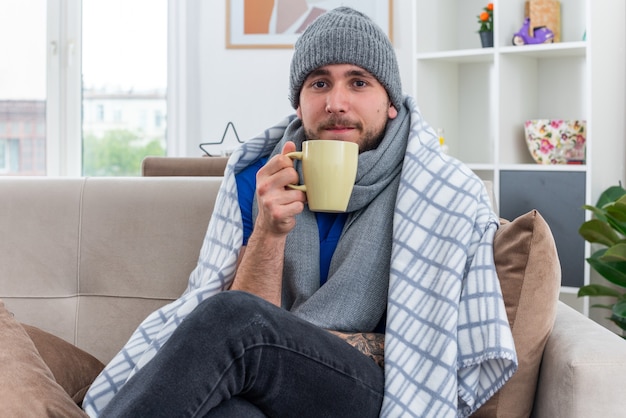 schwacher junger kranker Mann mit Schal und Wintermütze, der auf dem Sofa im Wohnzimmer sitzt und in eine Decke gehüllt ist, die eine Tasse Tee mit Blick auf die Vorderseite hält