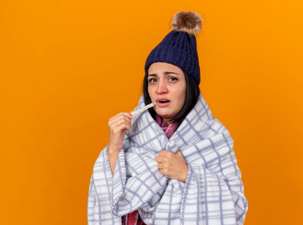 Schwaches junges kaukasisches krankes Mädchen, das Wintermütze und Schal trägt, die im Plaid eingewickelt sind, das Thermometer in den Mund ergreift, das Plaid lokalisiert auf orangefarbenem Hintergrund mit Kopienraum